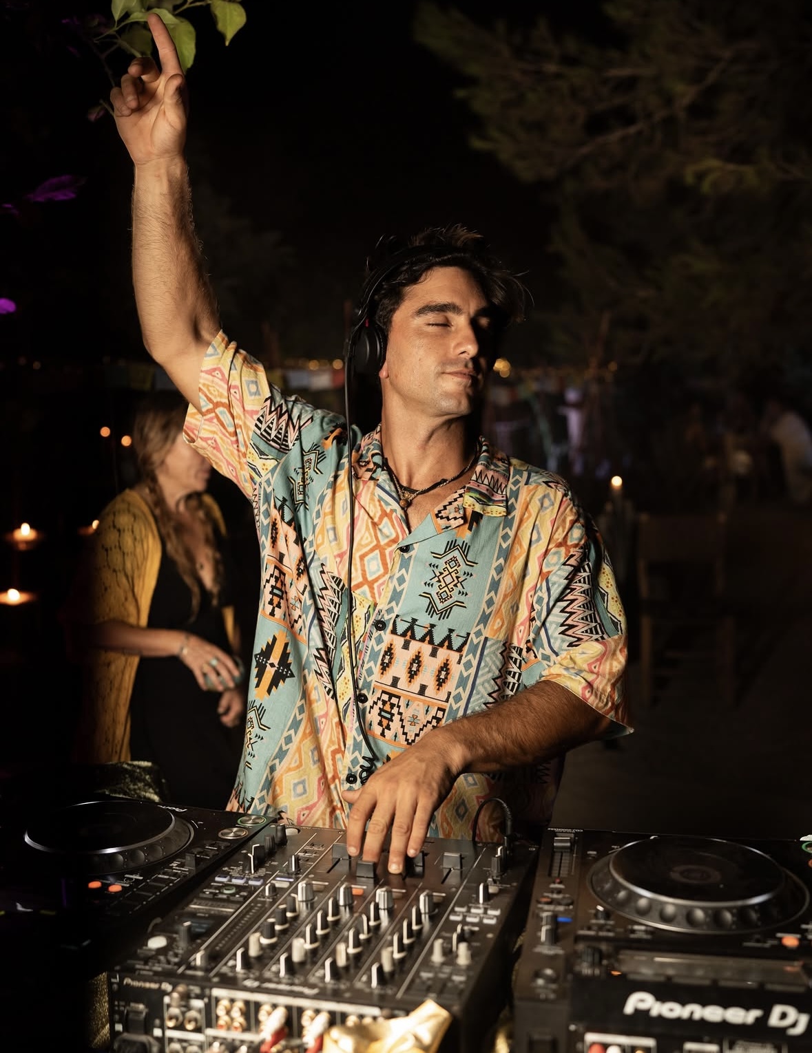 ORIAM behind the decks at night, hand raised to the trees